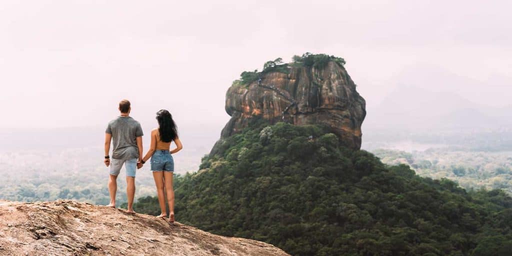 Ein Paar, das auf einem Felsen mit Blick auf einen riesigen Hügel und grüner Natur steht, symbolisiert Abenteuer und Freiheit im Zusammenhang mit Escort Service.