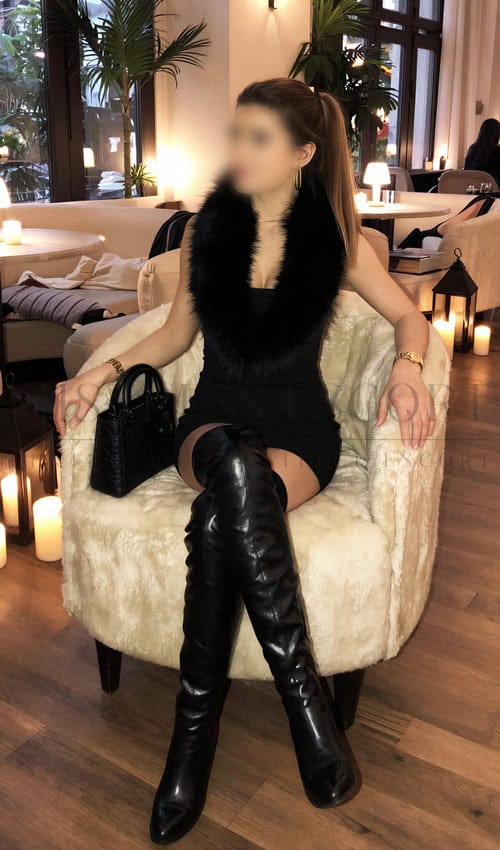 Attractive escort woman Annabelle in a stylish outfit with black over-the-knee boots and a fur scarf, sitting in a luxurious lounge area.