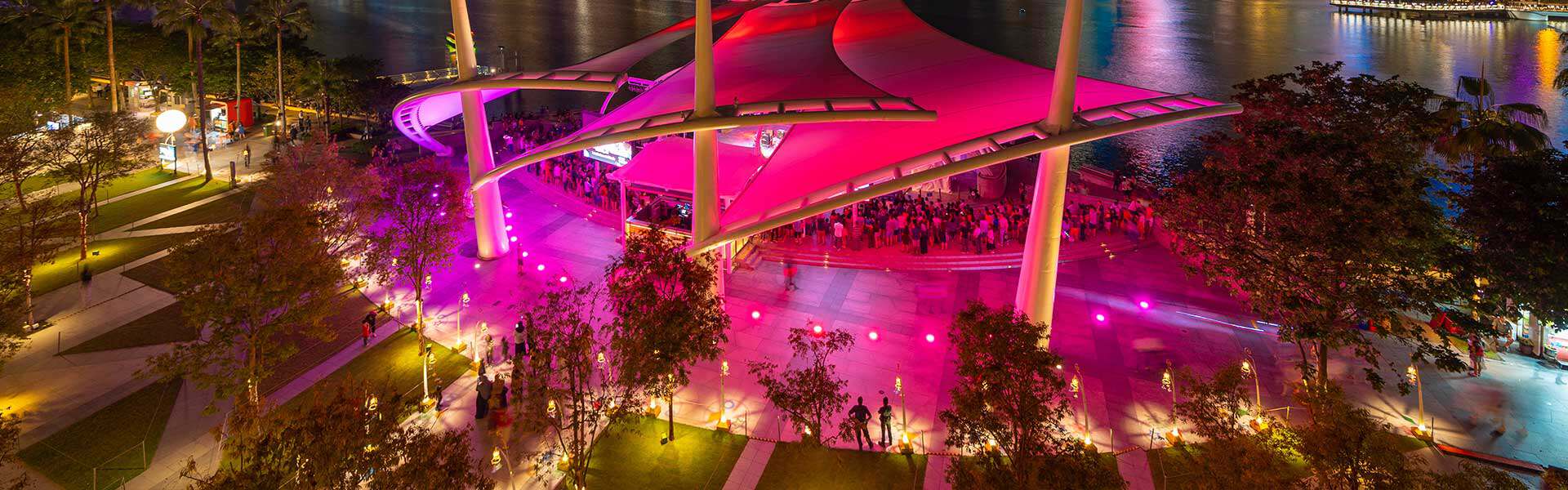 An elegant escort lady stands in a stylish, modern park at night, surrounded by colorful lights and reflective water surfaces, ideal for exclusive escort services.